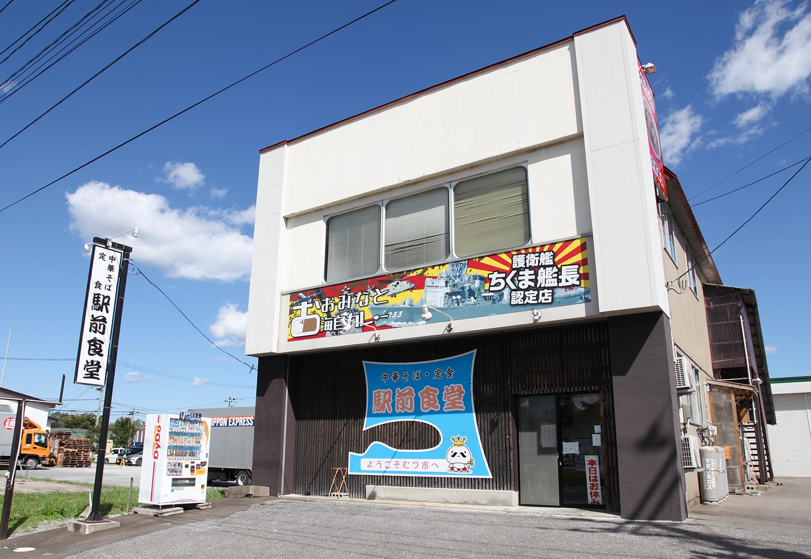 駅前食堂の外観：青空と『護衛艦ちくま艦長認定店』バナー、『ようこそむつ市へ』暖簾の入った店構え